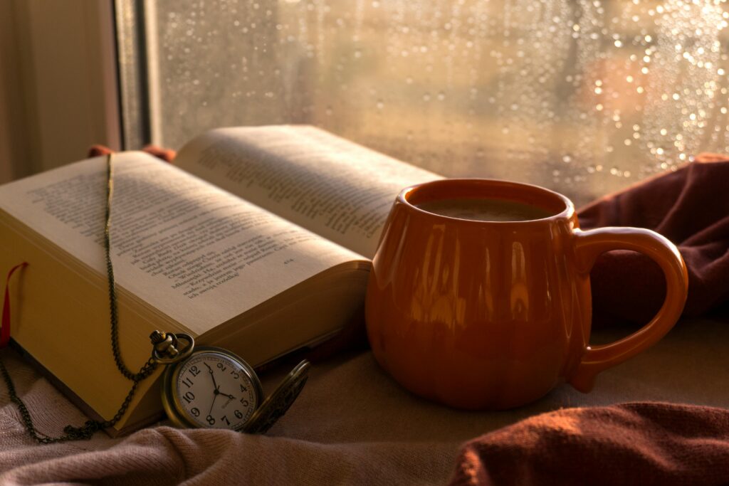 A warm autumn scene with a book, coffee, and pocket watch by the window on a rainy day.
