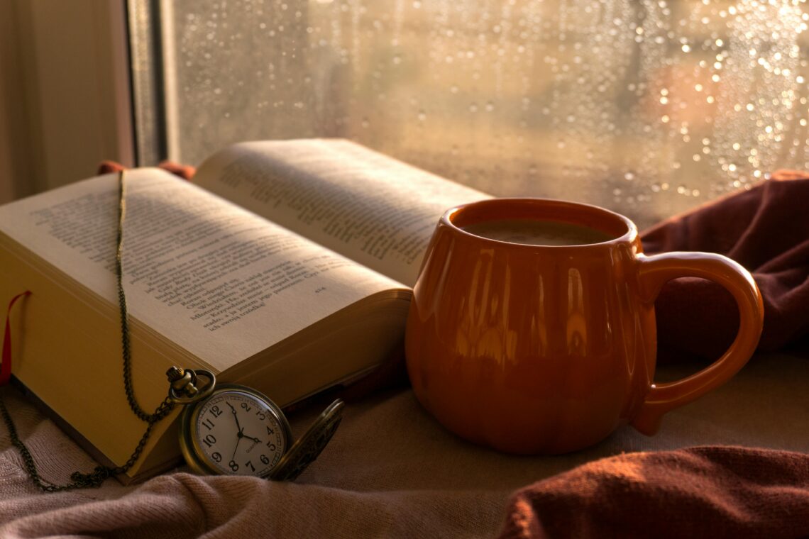 A warm autumn scene with a book, coffee, and pocket watch by the window on a rainy day.