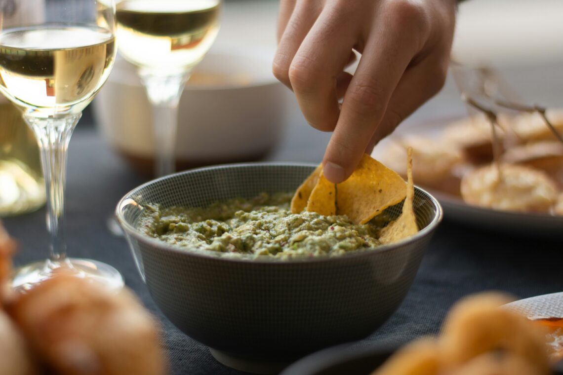 A hand dipping nachos into a bowl of fresh guacamole, accompanied by wine glasses.