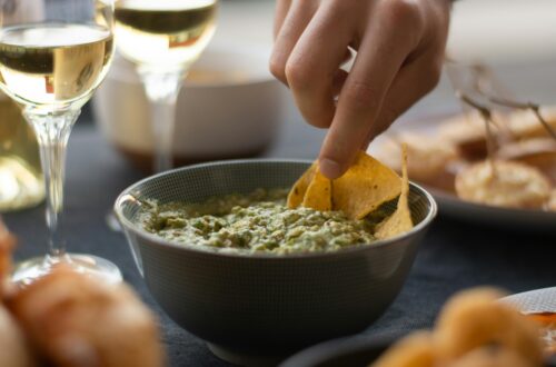 A hand dipping nachos into a bowl of fresh guacamole, accompanied by wine glasses.