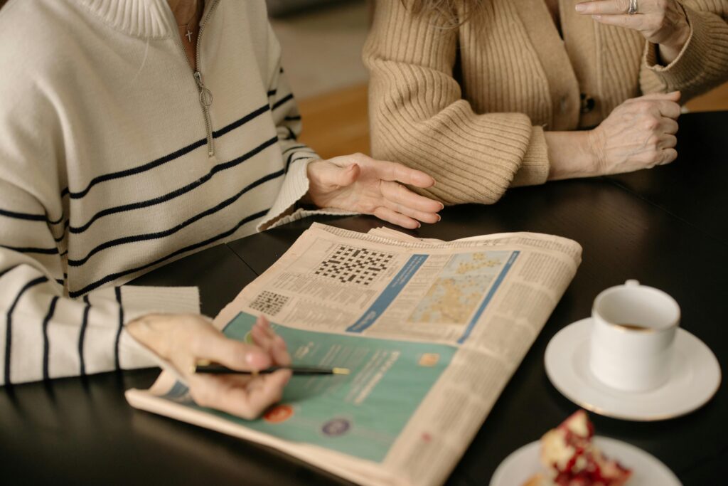 Two women discussing a newspaper crossword puzzle while enjoying coffee indoors.