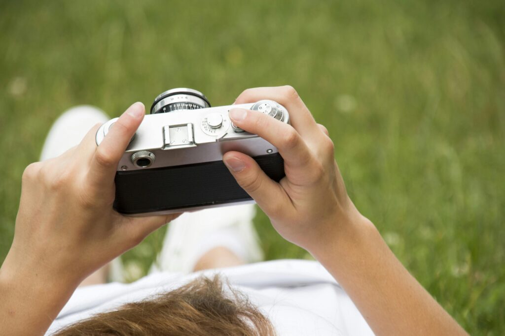 A pair of hands holding a vintage camera while lying on the grass, capturing an outdoor scene for his hobbies.
