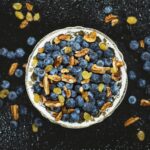 Top-down view of a bowl filled with fresh blueberries, pecans, and yellow raisins on a dark textured background.
