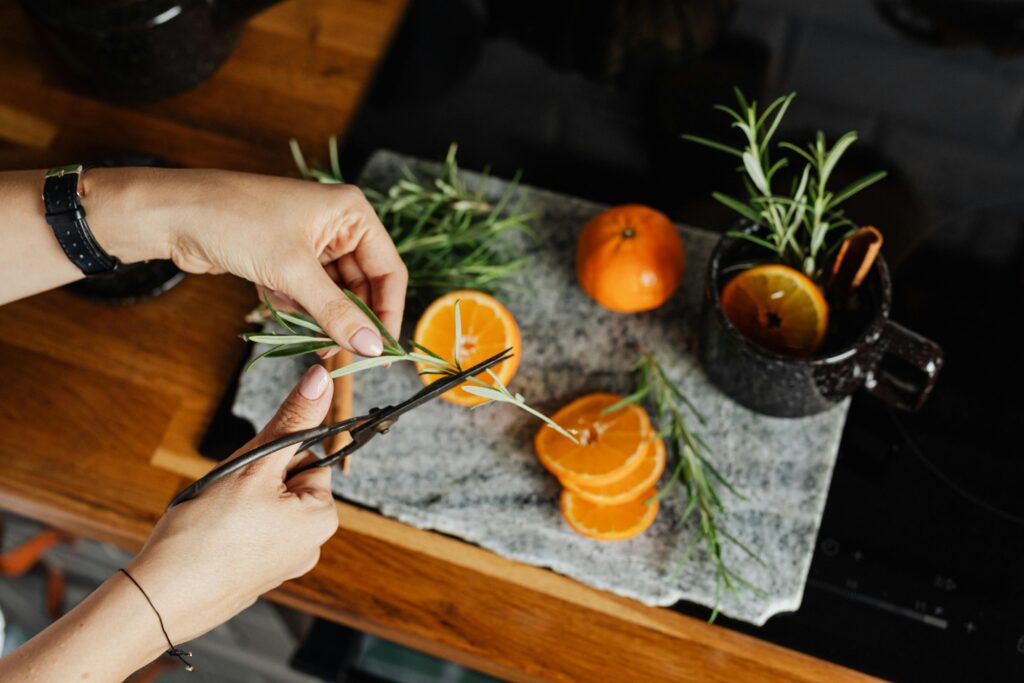 A close-up of hands cutting fresh rosemary next to sliced oranges on a kitchen counter top, ideal for a healthy classic tea. Photo by: by kaboompics | pexels